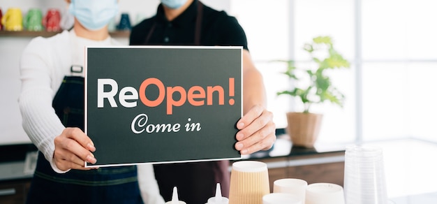 Young baristas couple wearing mask reopening their own cafe with a sign on a board.