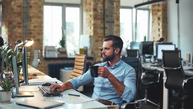 Young bearded businessman in formal wear holding cup of coffee and looking at computer 