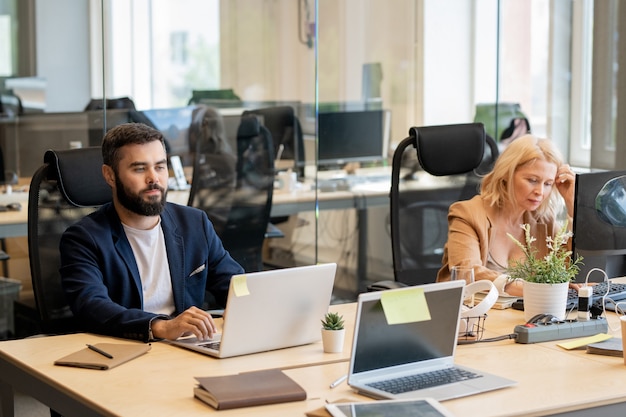 Young bearded businessman and mature blond female sitting by table in front of laptops in open space office and working individually