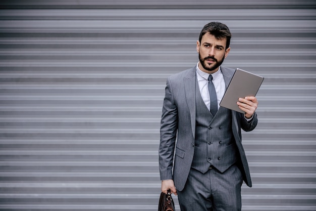 Young bearded businessman in suit holding tablet and briefcase.