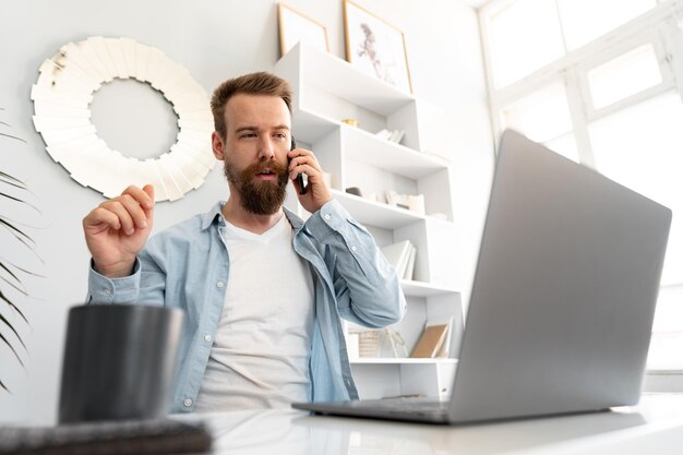 Photo young bearded businessman working from home sitting at the desk and talking on mobile phone