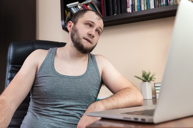  A young bearded guy in home clothes gray T-shirt, uses laptop