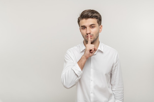 Young bearded handsome guy in white shirt makes gesture quietly isolated on white background