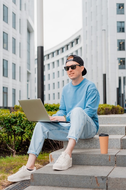 Young bearded hipster businessman stands on city street with cup of coffee and uses tablet computer.  Modern glass building man working blogging chatting online social media