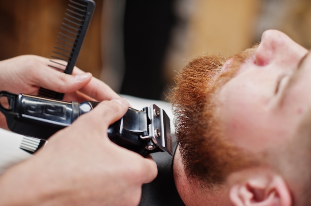 Young bearded man getting haircut by hairdresser while sitting in chair at barbershop. Barber soul.