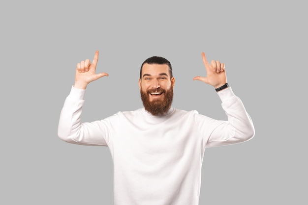 Young bearded man is pointing up with both hands indicating some copy space Studio shot over grey background