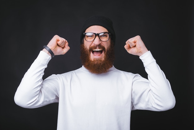 Young bearded man is screaming and holding both hand up near black wall.