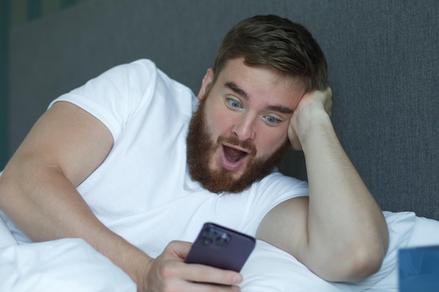 Young bearded man is using cell mobile phone at home in bed in bedroom looking at screen of