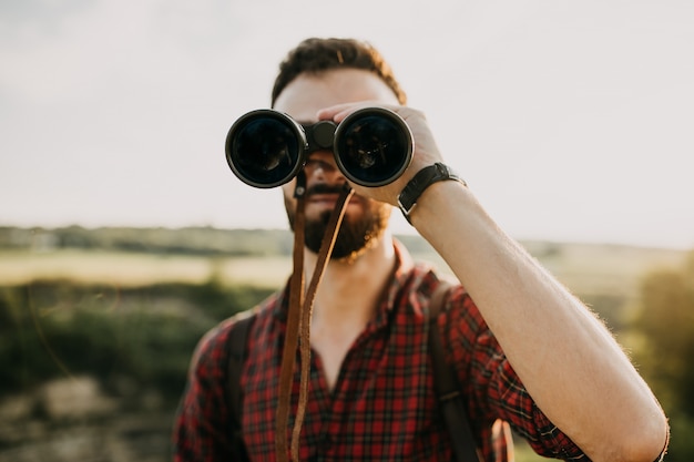 Young bearded man looking through binoculars, outdoors.