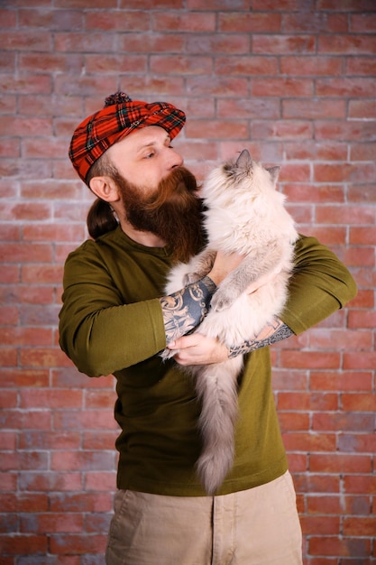 Young bearded man in plaid cap with fluffy cat on brick wall background