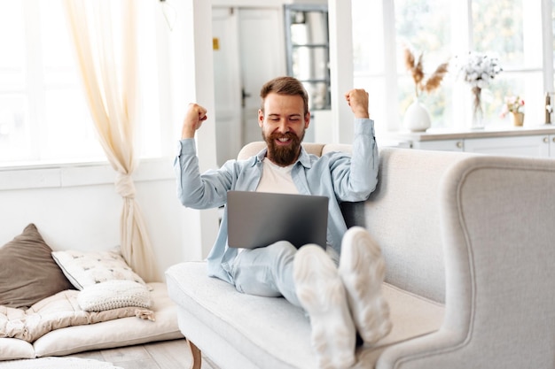 Young bearded man sitting on sofa at home using laptop computer