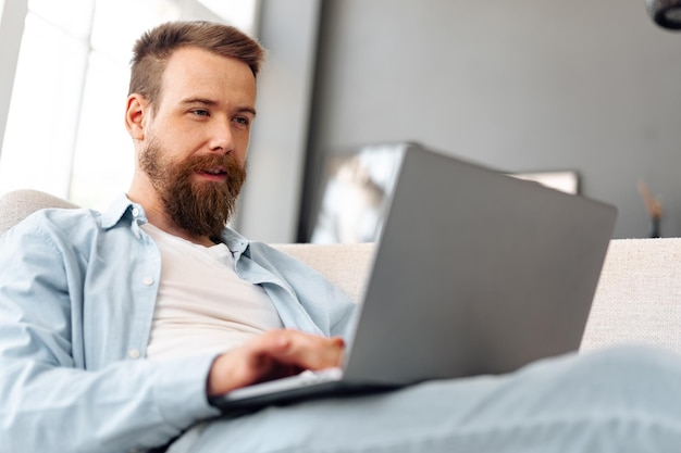 Young bearded man sitting on sofa at home using laptop computer