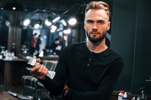 Photo young bearded man standing in barber shop and holding clipper