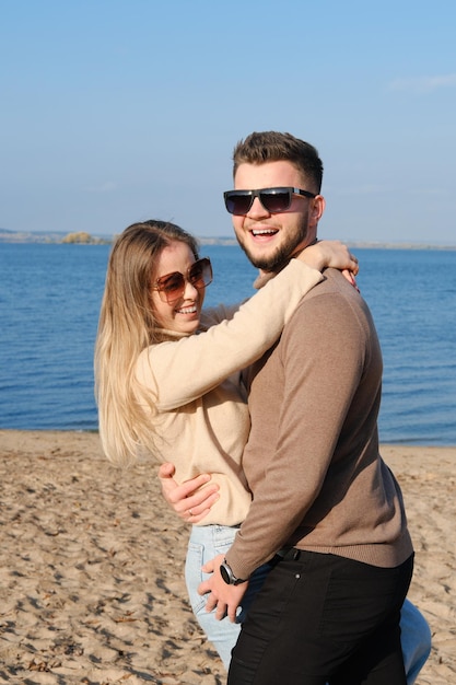 A young bearded man in sunglasses hugs a girl by the waist Couple having fun fooling around on the beach and looking at the camera