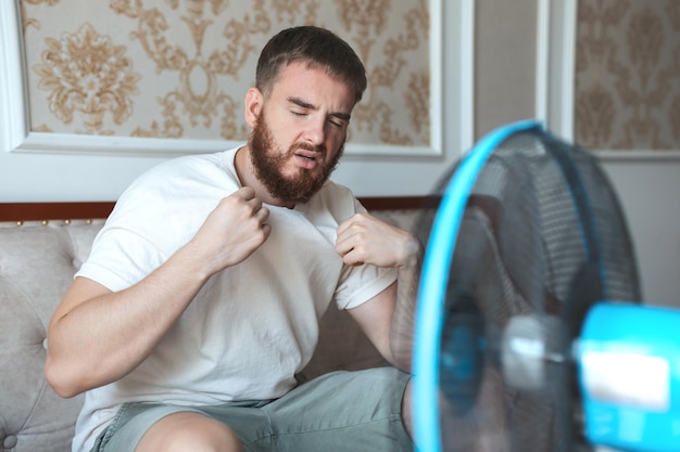 Young bearded man using electric fan at home sitting on couch cooling off during hot weather