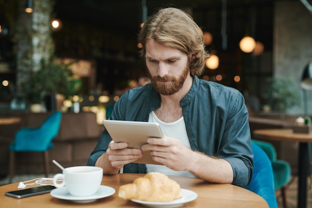 Young bearded manager sitting at table with coffee cup and croissant and recording voice message for