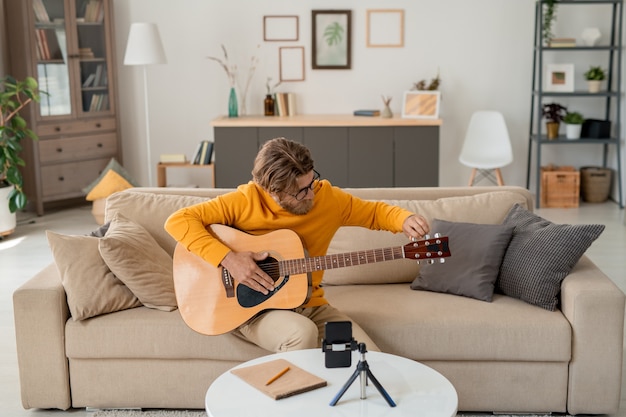 Young bearded musician or music teacher in casualwear sitting on couch in living-room and playing guitar in front of smartphone camera