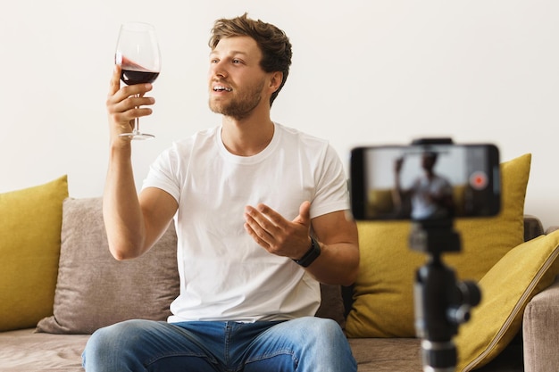 Young bearded sommelier blogger sitting on sofa in front of a camera is looking at a glass of red wine in a brightly lit room