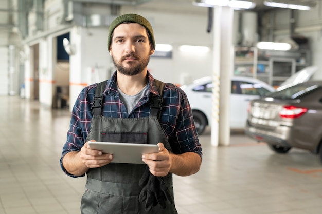 Young bearded worker of contemporary car service using digital tablet while asking online questions of clients in working environment