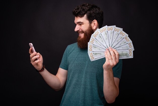 Young bearding man betting on his phone is holding a bunch of money in his hand.