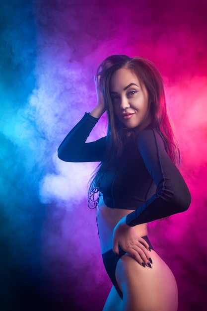 Young beatiful woman in black underwear posing against a background of blue and pink smoke from a vape on a black isolated background