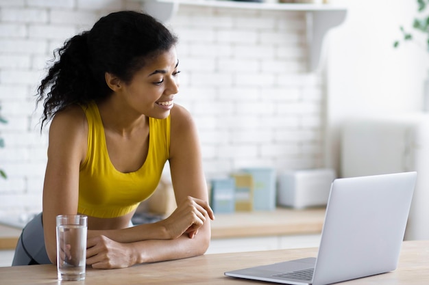 Young beautiful African American woman fitness trainer using laptop by video call, provides an online consultation on healthy eating and fitness standing in the home kitchen