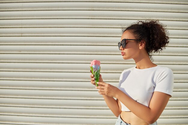 A young beautiful AfricanAmerican girl in a white tshirt and light jeans eats ice cream against a light wall on a Sunny day