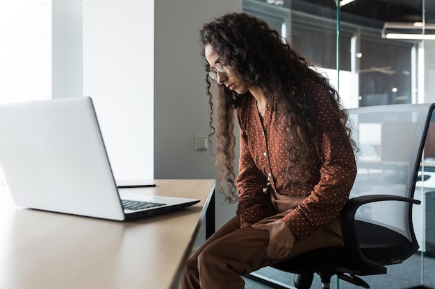 Young beautiful arab woman working in modern office with laptop curly business woman has severe pain