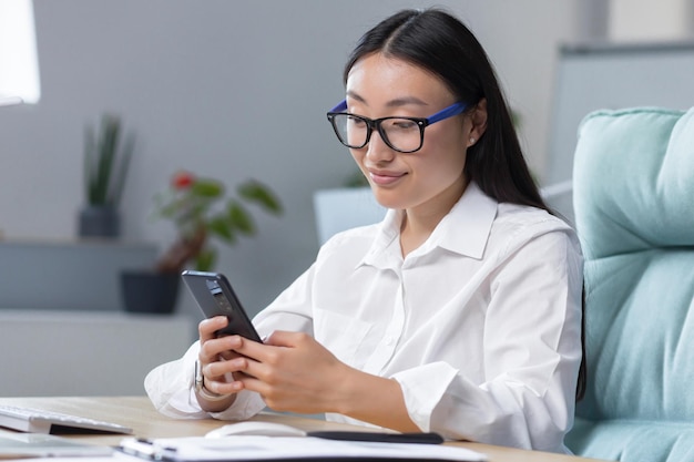 Young beautiful asian business woman works in modern office uses phone smiles reads good news chats