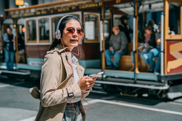 young beautiful asian chinese woman commuter in city street with headphones listening to music from cellphone app online. relax technology lifestyle concept. cable car in san francisco in background