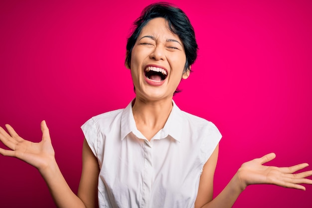 Young beautiful asian girl wearing casual summer shirt standing over isolated pink background celebrating mad and crazy for success with arms raised and closed eyes screaming excited Winner concept