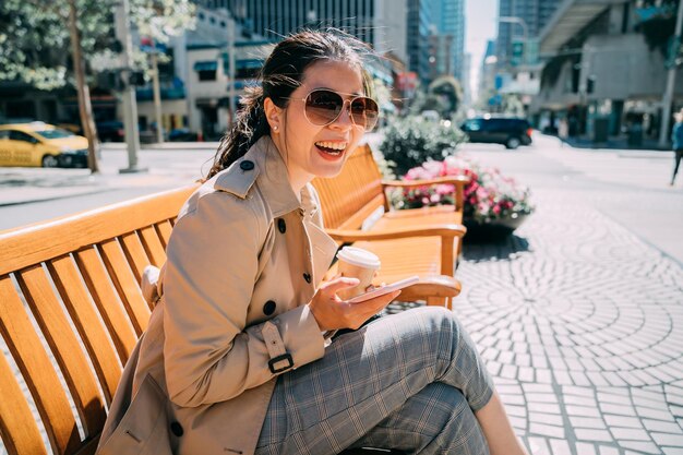 Young beautiful asian korean girl sitting on wooden bench holding paper cup of coffee and cellphone. confident positive woman in fashion sunglasses cheerful laughing and using mobile phone outdoor