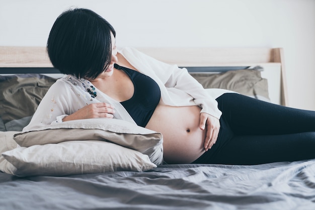 Young beautiful Asian pregnant woman lying in bed and touching her belly at home