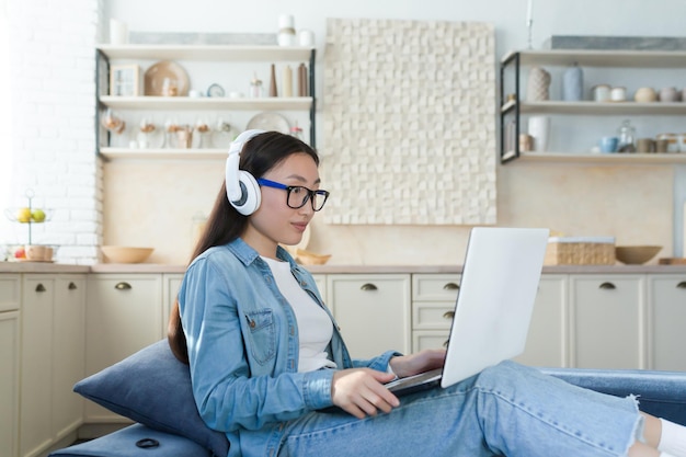 Young beautiful asian teenage girl in headphones resting on sofa at home sitting on the couch