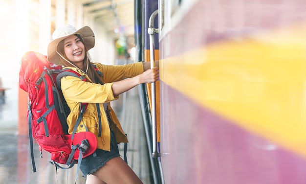 Young beautiful asian traveler with backpack getting on a train at a platform of suburban railway station, smiling and looking at front