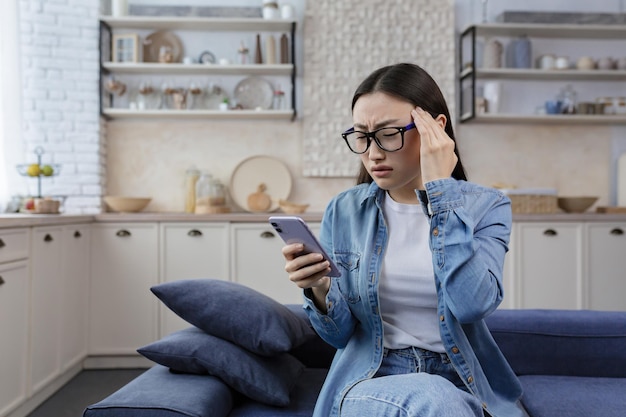 Young beautiful asian upset woman sitting at home in living room on sofa wearing glasses reading