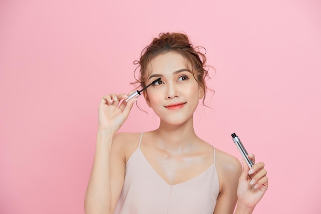 Young beautiful Asian woman applying black mascara on eyelashes with makeup brush over pink background