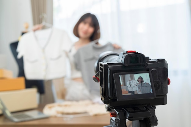 Young and beautiful Asian woman blogger showing clothes in front of smartphone camera while recording vlog video