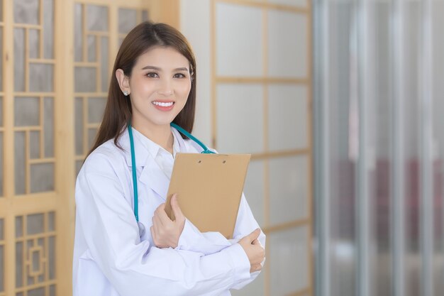 Young beautiful  Asian woman doctor Standing with arms crossed happy and smile in hospital. Wearing a white robe and stethoscope