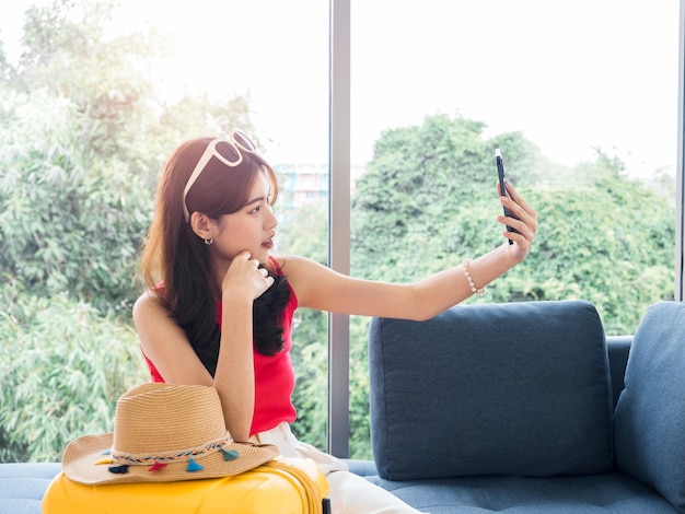 Young beautiful Asian woman holding smart mobile phone and make selfie by camera Happy Asian female wearing sunglasses and beach hat near the yellow suitcase sit on couch travel summer vacation