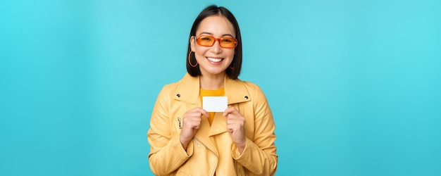 Young beautiful asian woman showing credit card smiling choosing bank standing over blue background Copy space