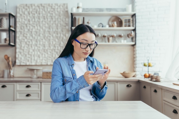 Young beautiful asian woman sitting in kitchen at home and using mobile phone scrolls through the