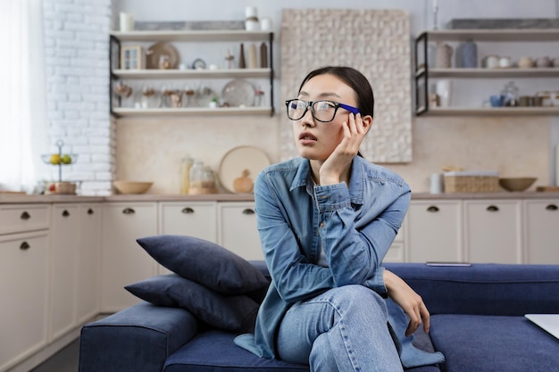 Young beautiful asian woman thinking in glasses sitting on sofa at home in living room woman sad in