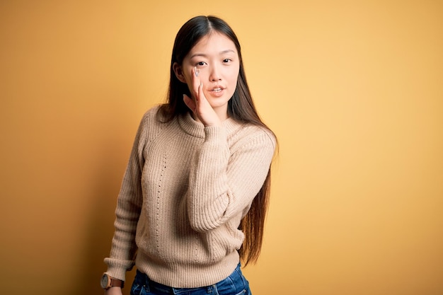 Young beautiful asian woman wearing casual sweater over yellow isolated background hand on mouth telling secret rumor whispering malicious talk conversation