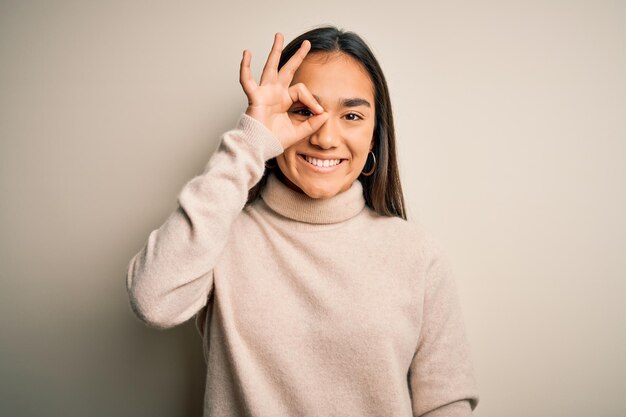 Young beautiful asian woman wearing casual turtleneck sweater over white background doing ok gesture with hand smiling eye looking through fingers with happy face