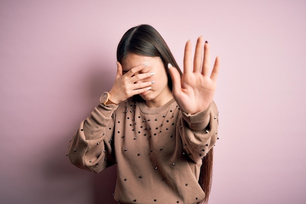 Young beautiful asian woman wearing fashion and elegant sweater over pink background covering eyes