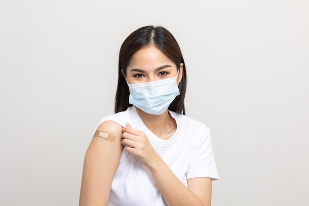 Young beautiful asian woman wearing mask and getting a vaccine protection the coronavirus. Happy female showing arm with bandage after receiving vaccination on isolated white background.