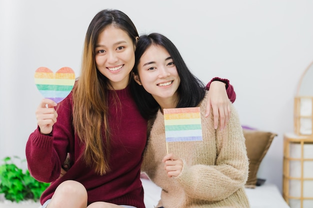 Young beautiful Asian women lesbian couple lover holding LGBT flag and community color heart shape in living room at home with smiling faceConcept of LGBT sexuality with happy lifestyle together