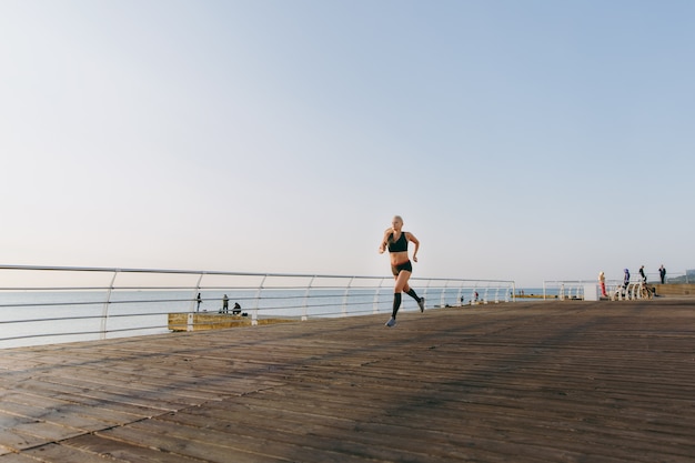 The young beautiful athletic girl with long blond hair in black clothes running at sunrise over the sea