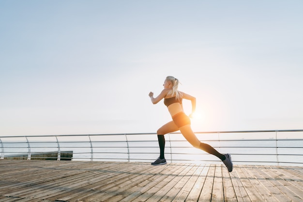 The young beautiful athletic girl with long blond hair in black clothes running at sunrise over the sea